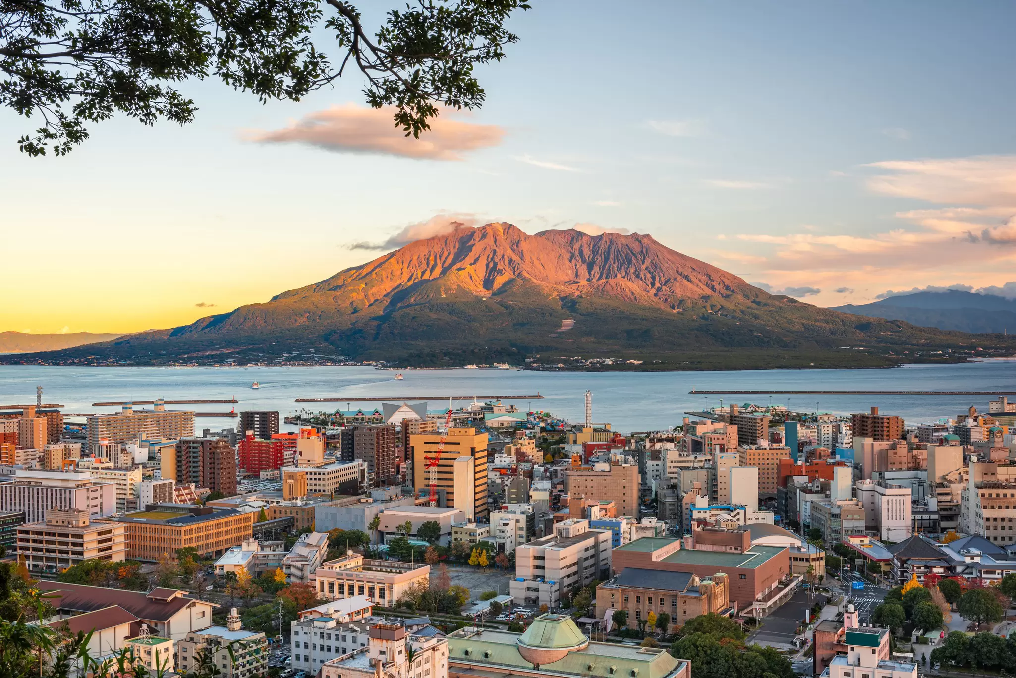 A volcano lit by sunrise is across the water from a city.
