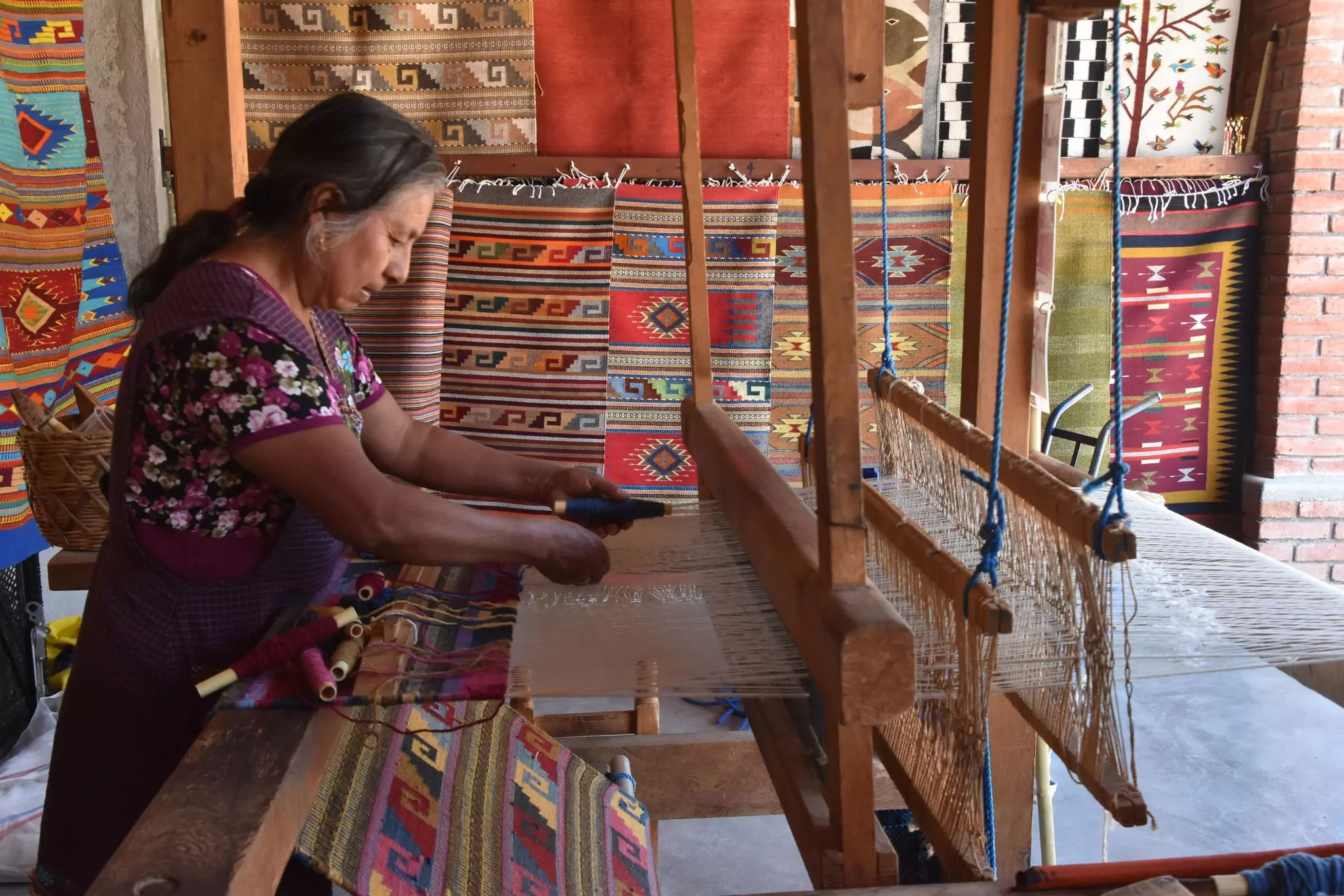 An older woman works a traditional loom, with hand-woven textiles hanging on the walls surrounding her.