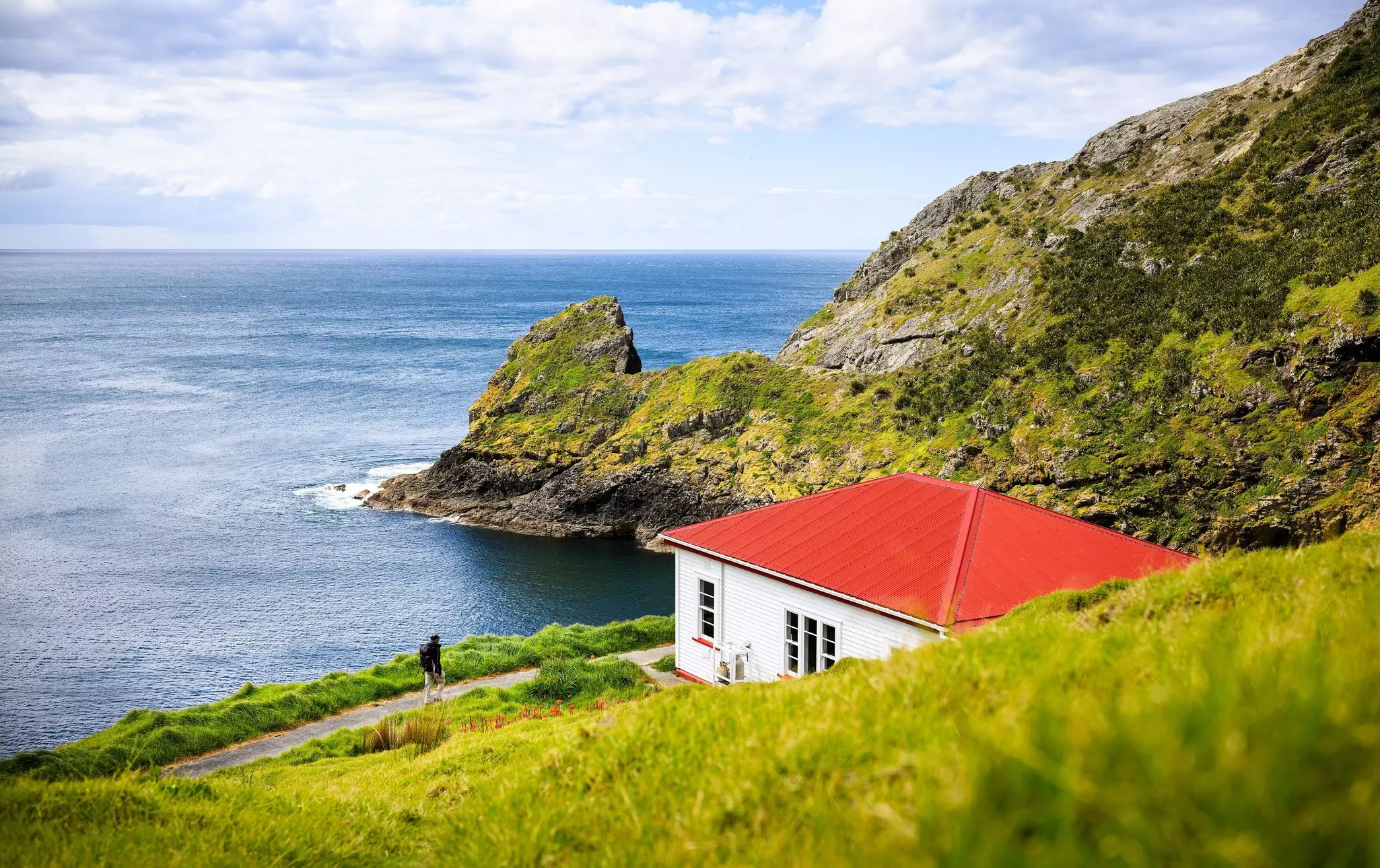 A hiker following a coastal path approaches a red-roofed hut tucked into the edge of a hillside.