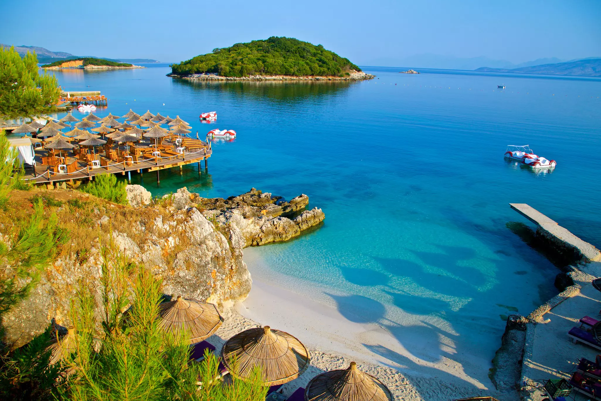 A wooden pier on the right, a collection of tables on the left, and a beach in the middle stretches into shallow blue waters off the coast of Ksamil beach, with a small island, covered in trees, in the distance.