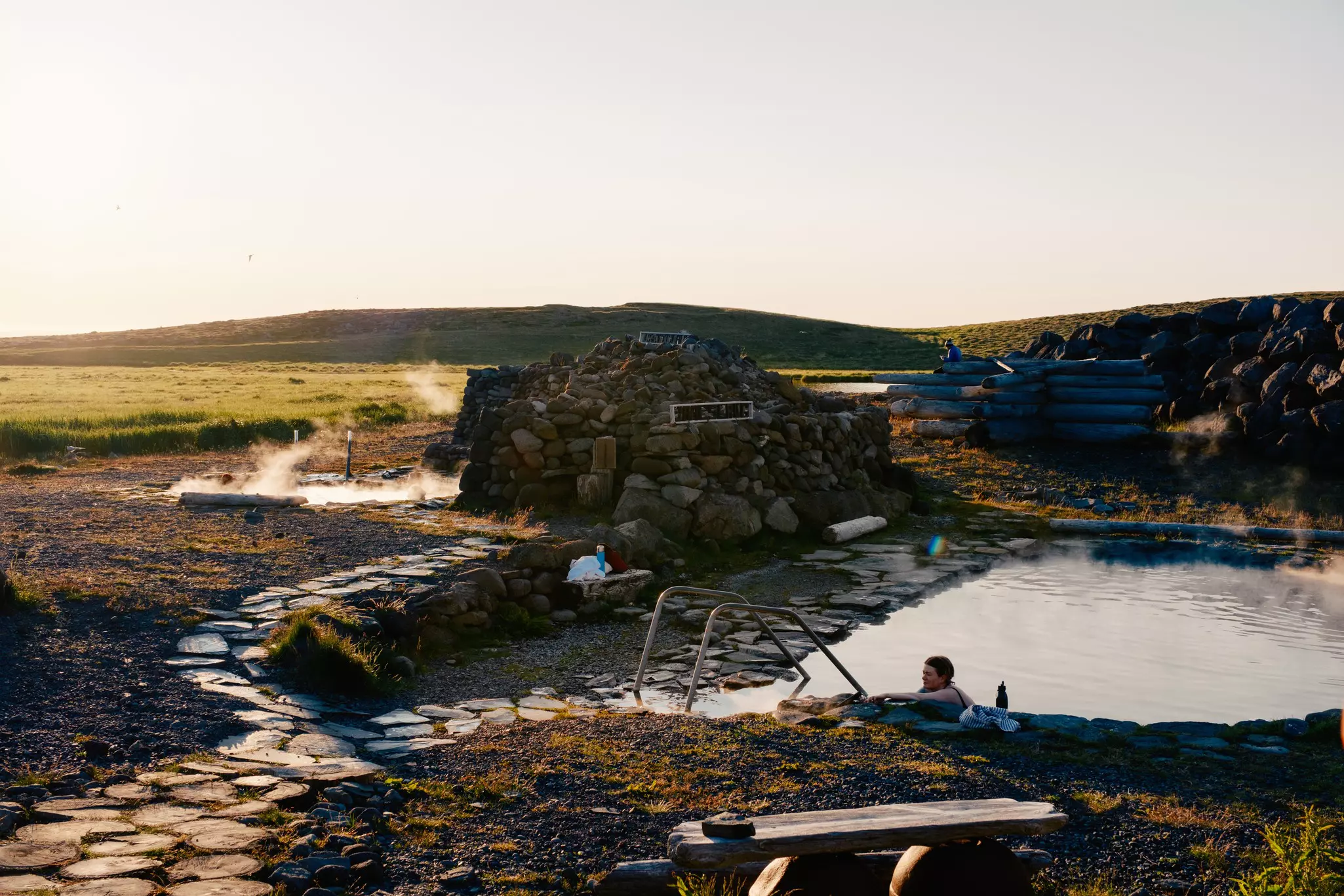 A woman raches out of a geothermal pool surrounded by rocks with steam vents and a grassy hillside in the distance on a mostly sunny day.