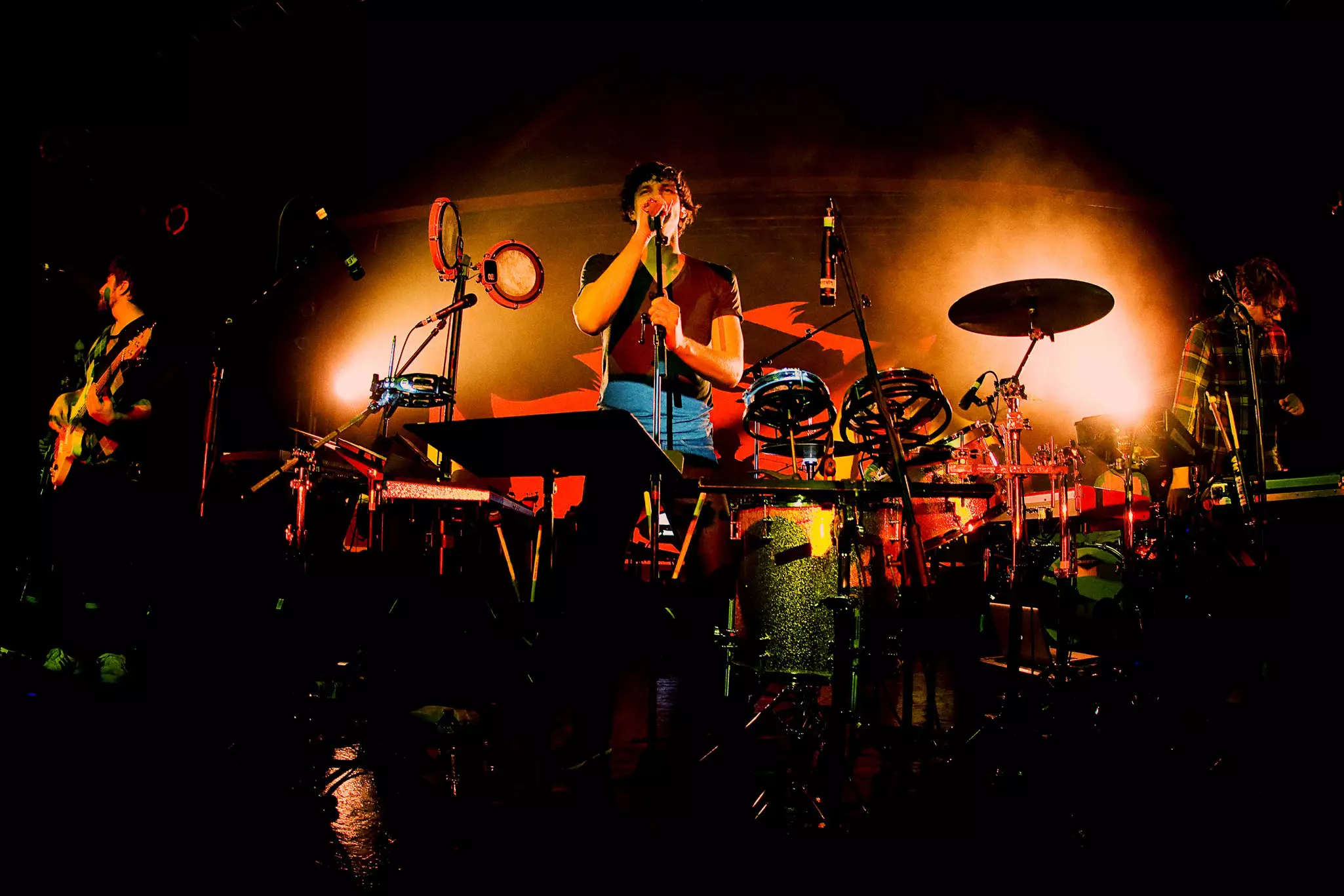 Three musicians performing on stage with the one in the center singing into a microphone under an orange spotlight.