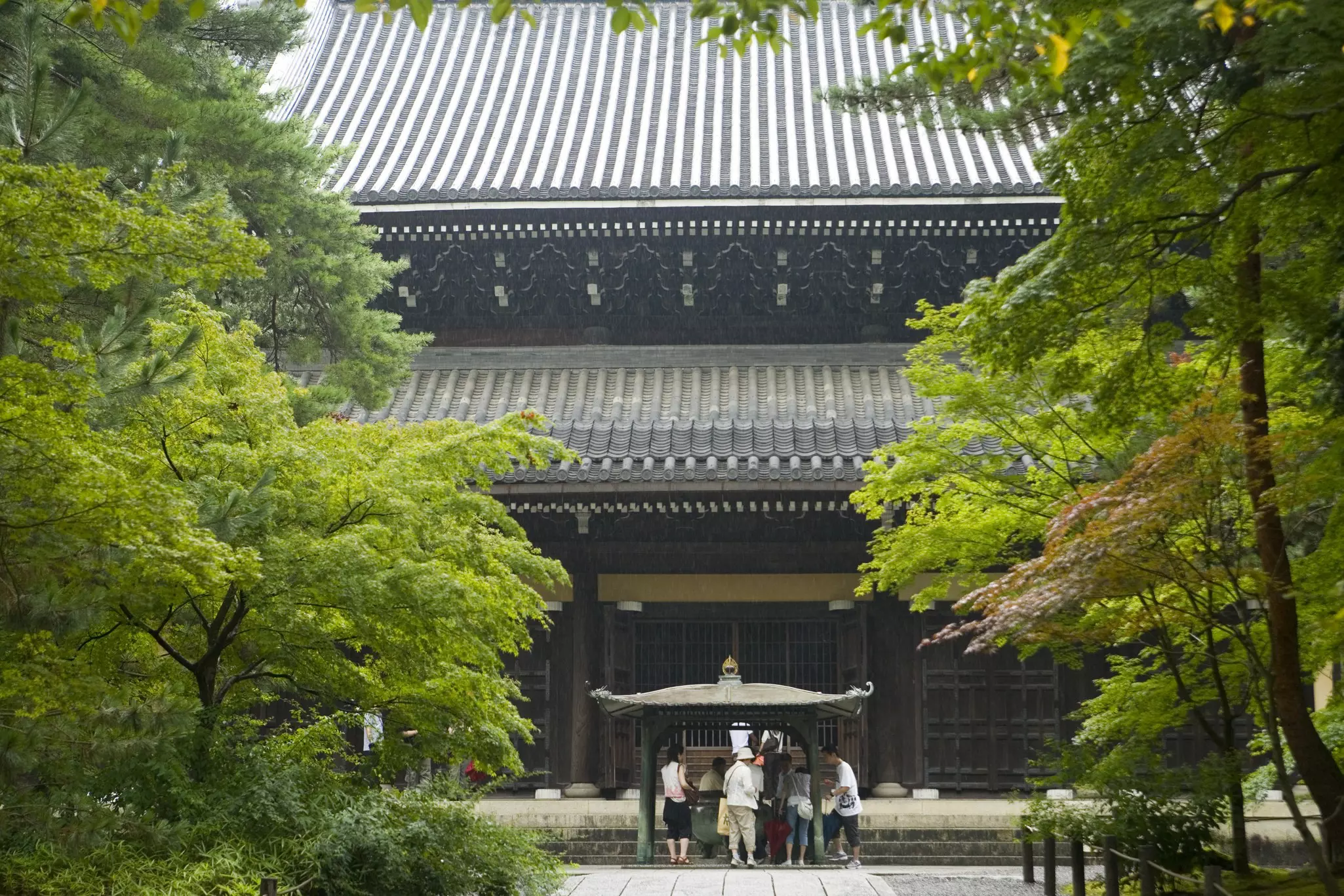 People stand in front of the pavilion of a large temple surrounded by verdant trees.