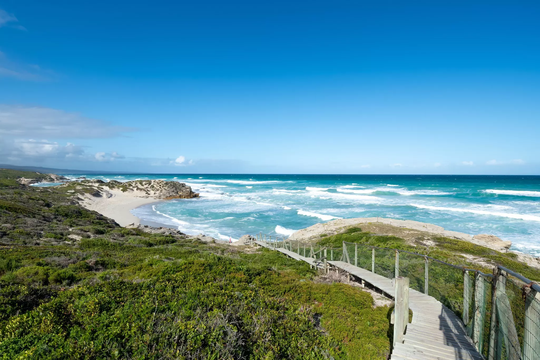 A boardwalk over shrubs and grassland leads down to a sandy beach being pounded by the surf.