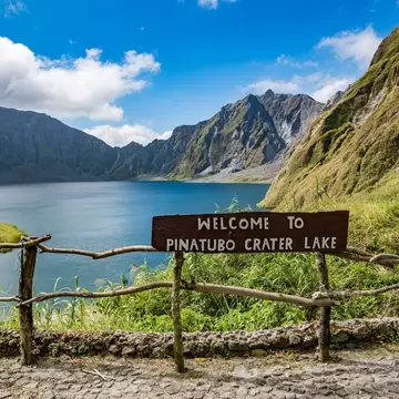 Wooden sign on a rustic railing overlooks a blue lake enclosed by steep, green-streaked crater walls