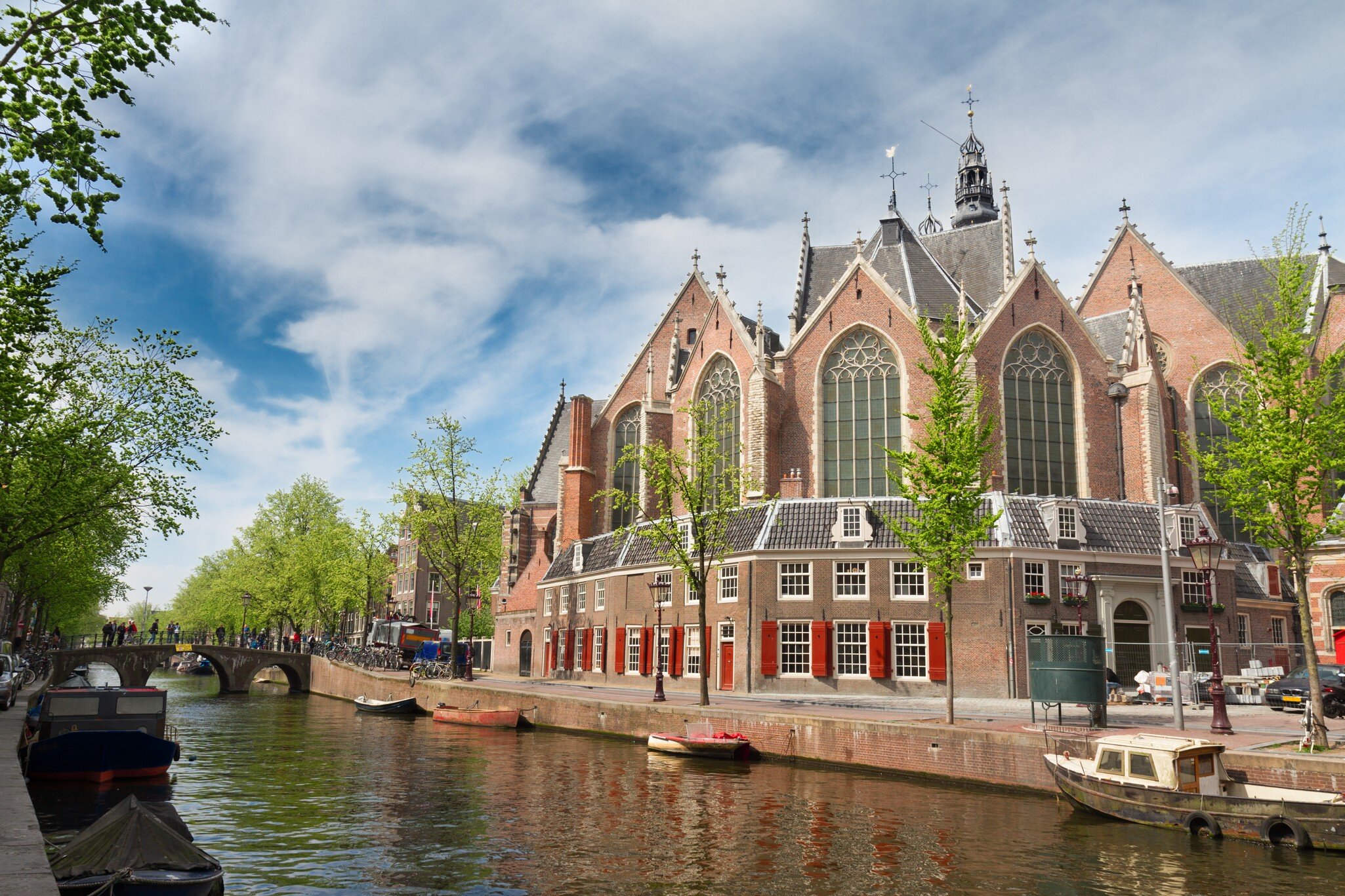Brick Gothic church with tall arched windows behind houses with red shutters, small boats moored on the canal.