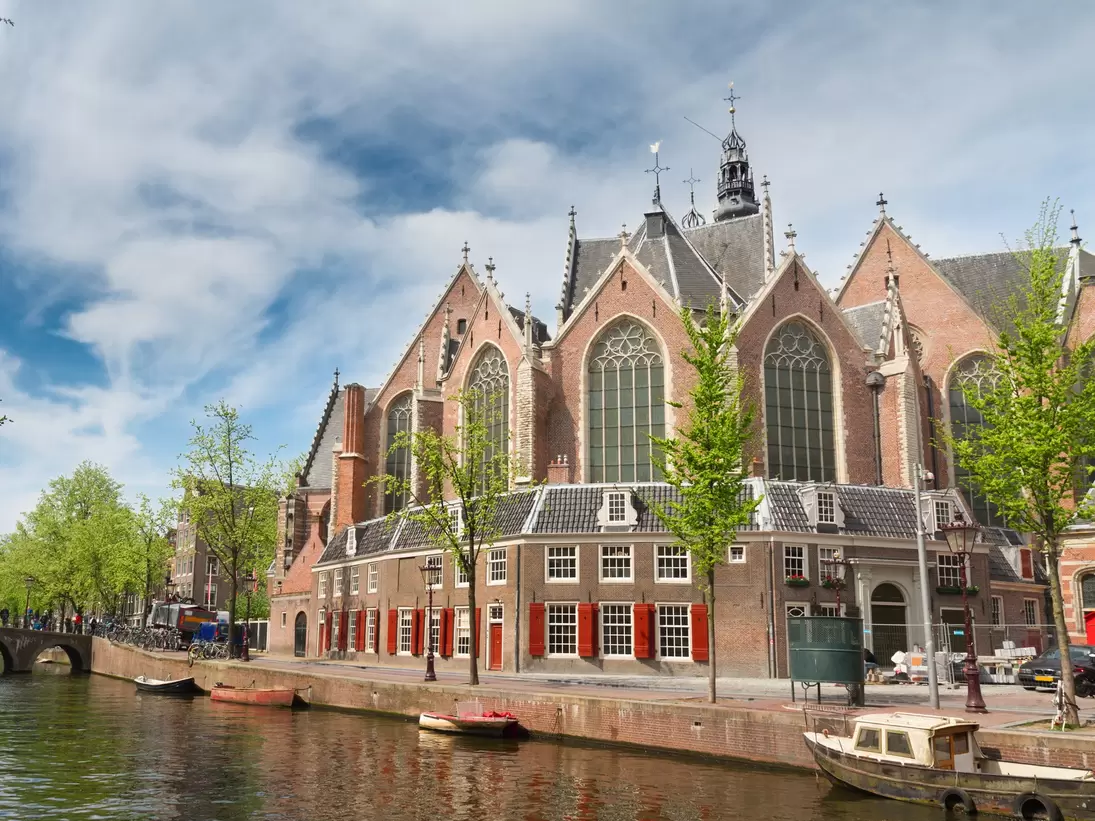 Brick Gothic church with tall arched windows behind houses with red shutters, small boats moored on the canal.