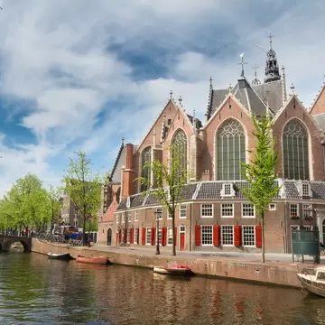 Brick Gothic church with tall arched windows behind houses with red shutters, small boats moored on the canal.