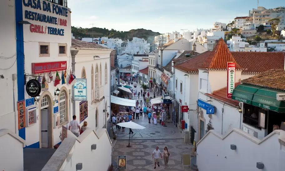 Albufeira old town: crowds on a cobbled street past whitewashed bars incl. Casa da Fonte and O Tunel, hillside homes above