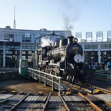 A historic steam locomotive is on display at a roundhouse as part of a museum exhibit.