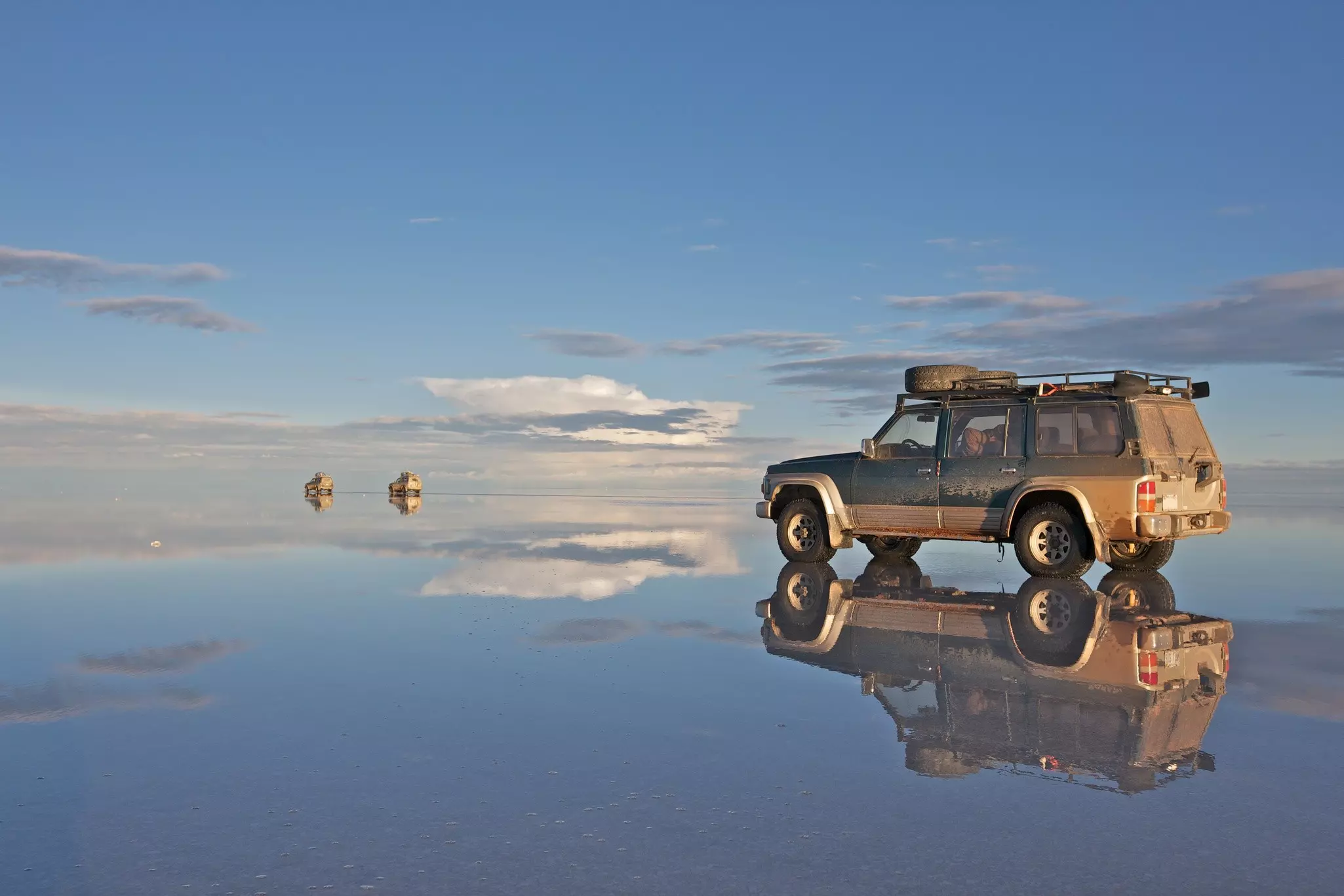 A 4WD stands in shallow water creating a near-perfect reflection. It is in an extensive flat area that seems to stretch endlessly