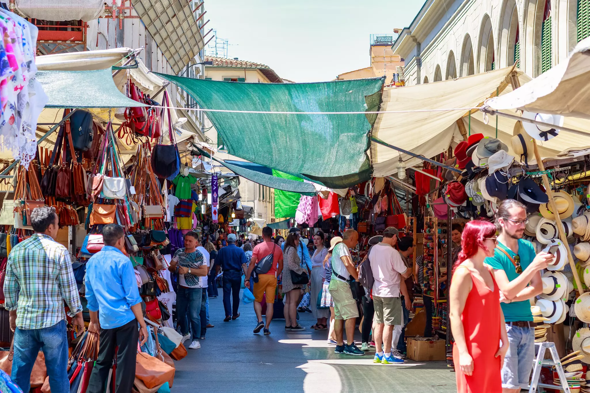 A busy outdoor market; shoppers look at hats, leather bags and other items under fabric shades.