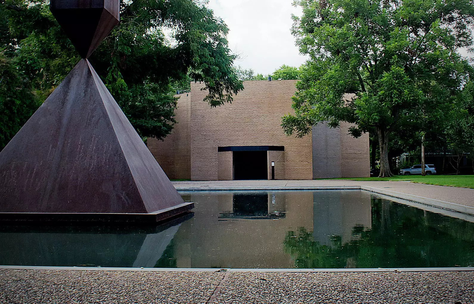 A peaceful pool with a large triangular sculpture in the center outside a square brick building.