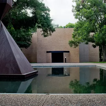 A peaceful pool with a large triangular sculpture in the center outside a square brick building.