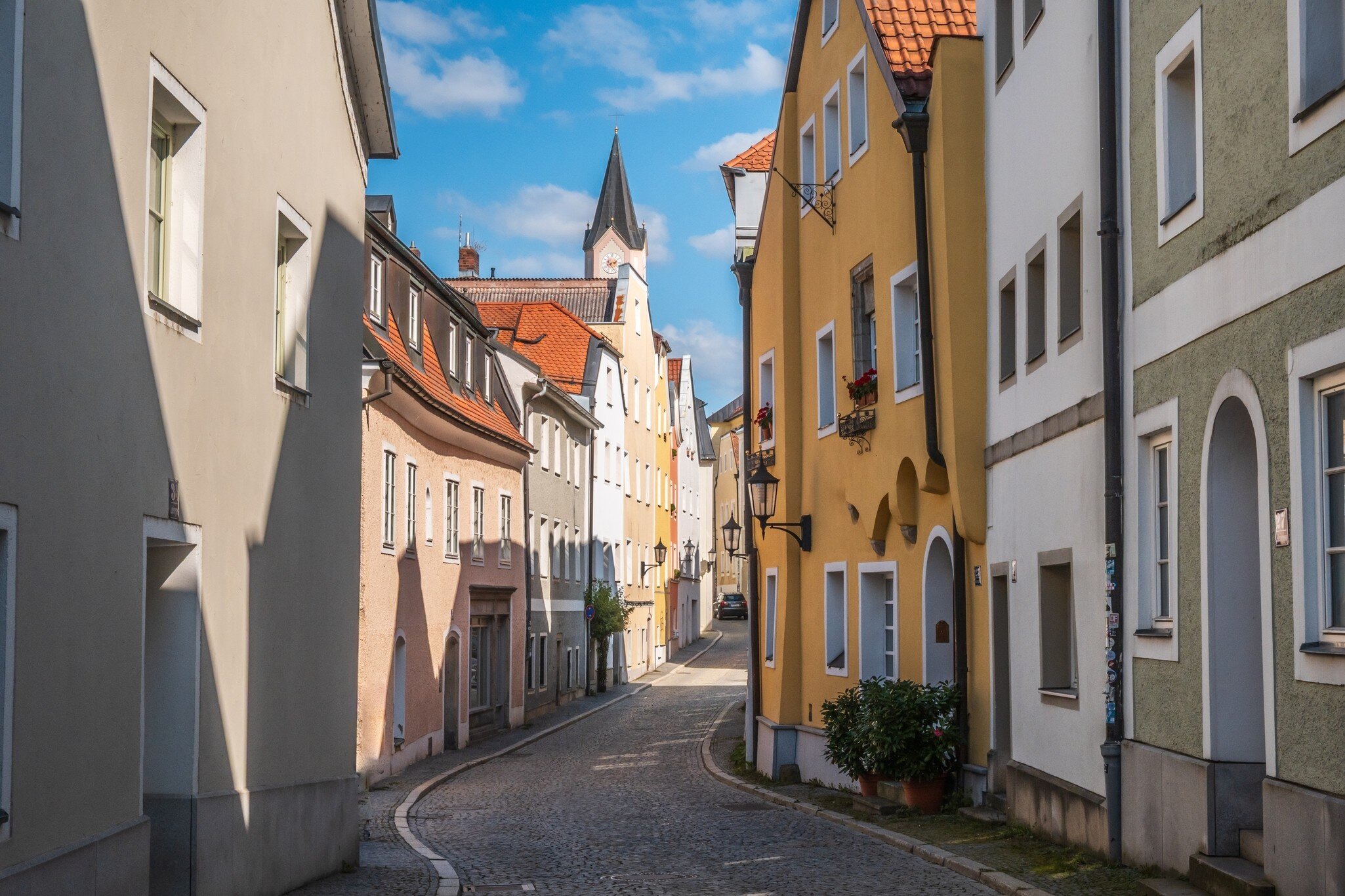 Pastel townhouses line a curving cobbled street in Passau that leads to a church spire under a blue sky