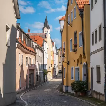 Pastel townhouses line a curving cobbled street in Passau that leads to a church spire under a blue sky