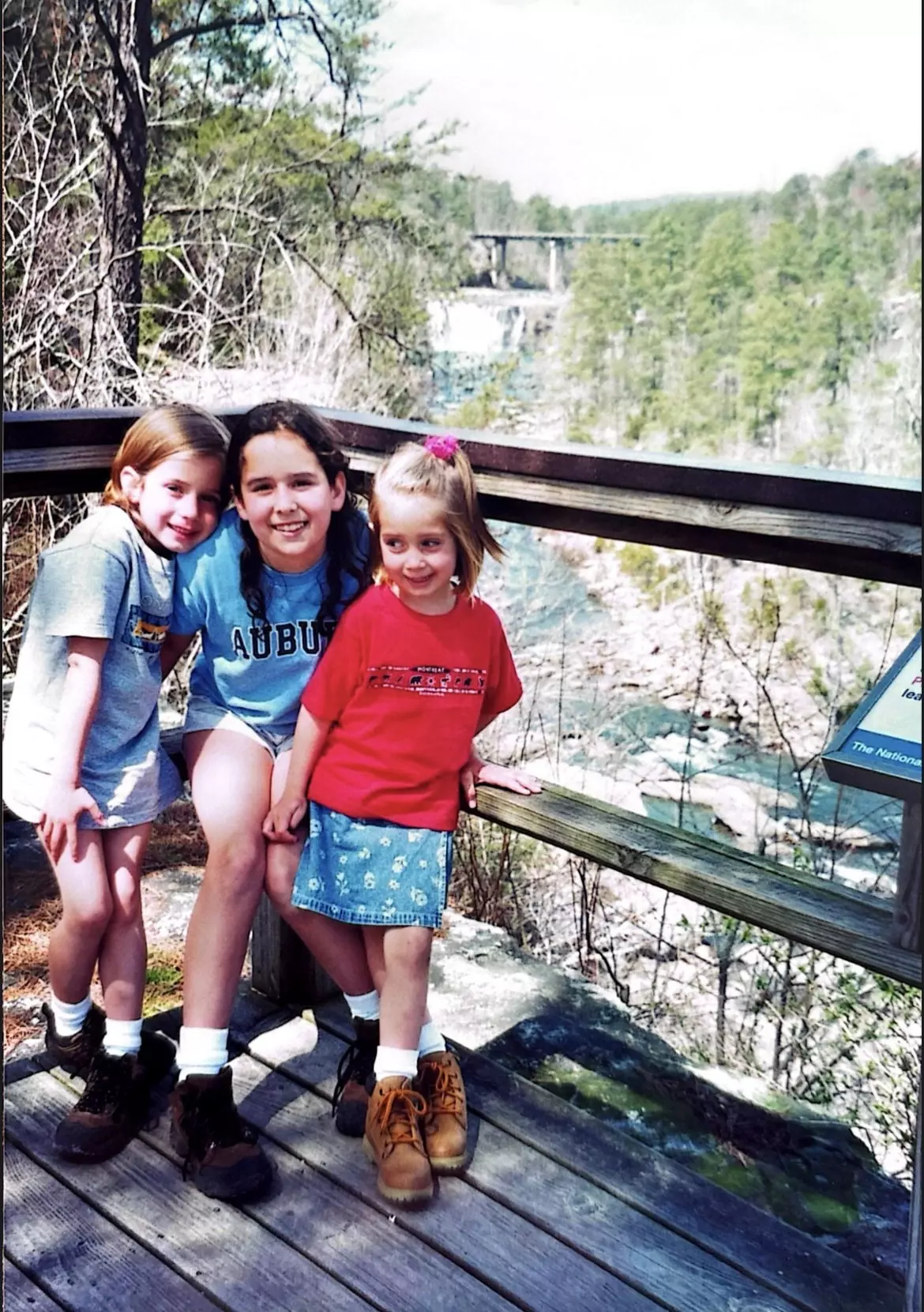 Three children posing for a photo against a wooden railing on a sunny day.