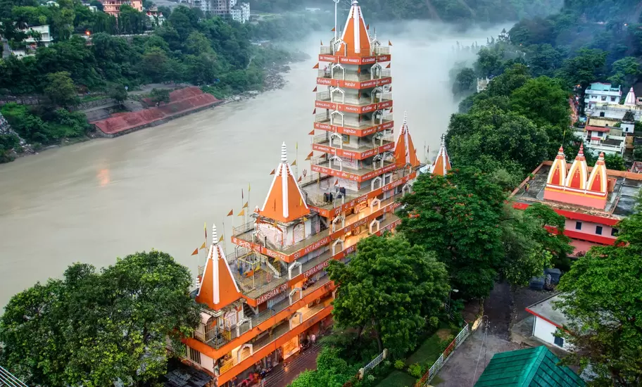 Orange-and-white multi-tiered Trimbakeshwar temple beside the misty Ganges in Rishikesh, forested hills behind