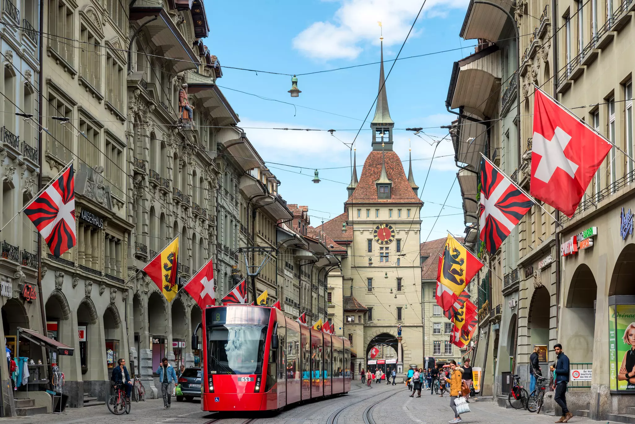 A red tram runs down a street with stone buildings on either side; multicolored flags hang from the buildings.