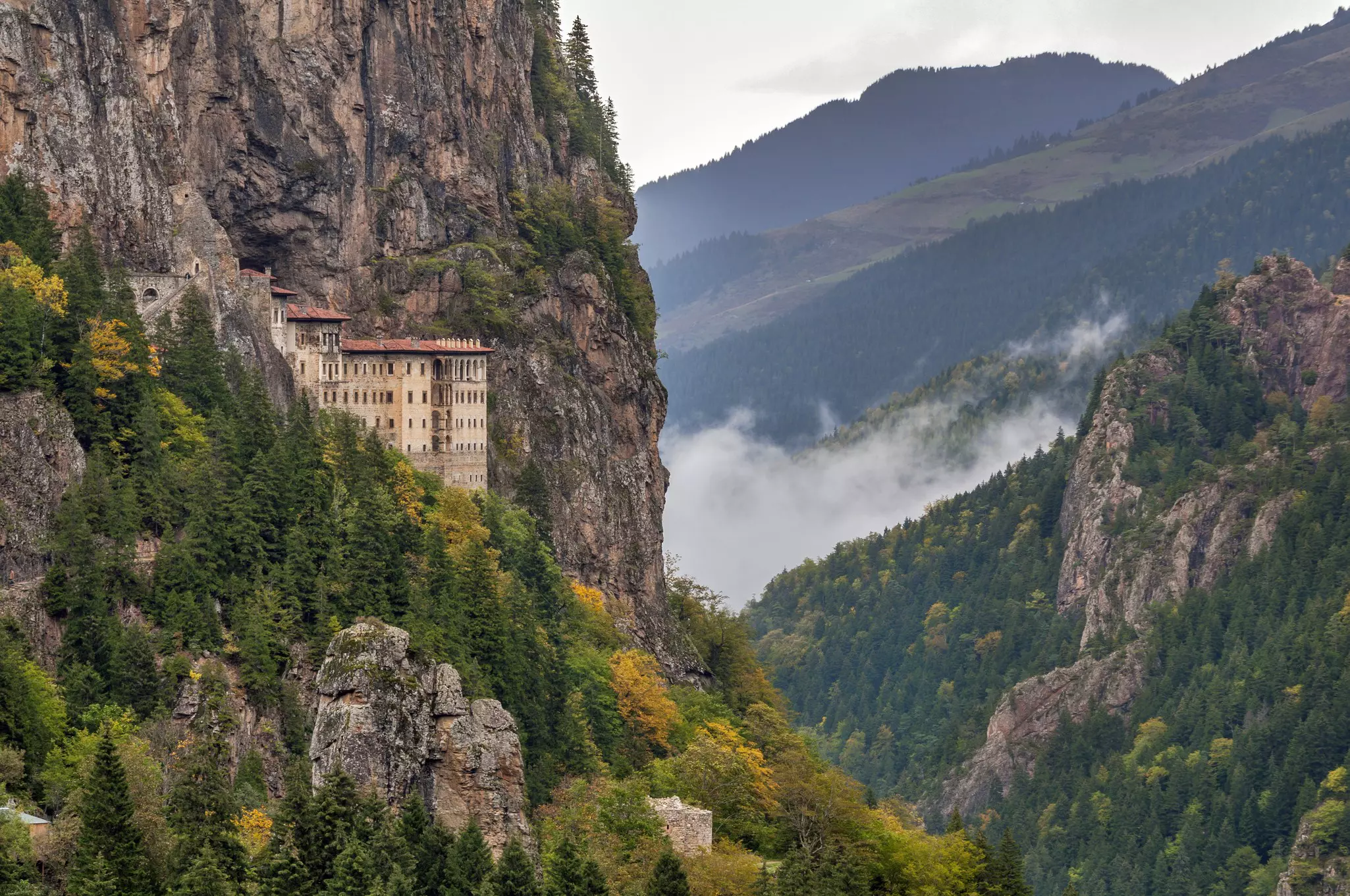 A building sits on the edge of a sheer rocky cliff in Türkiye; there is a cloud in a deep valley in the background.