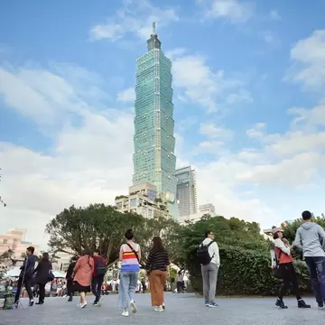 People walking around at the base of the light green Taipei 101 tower.