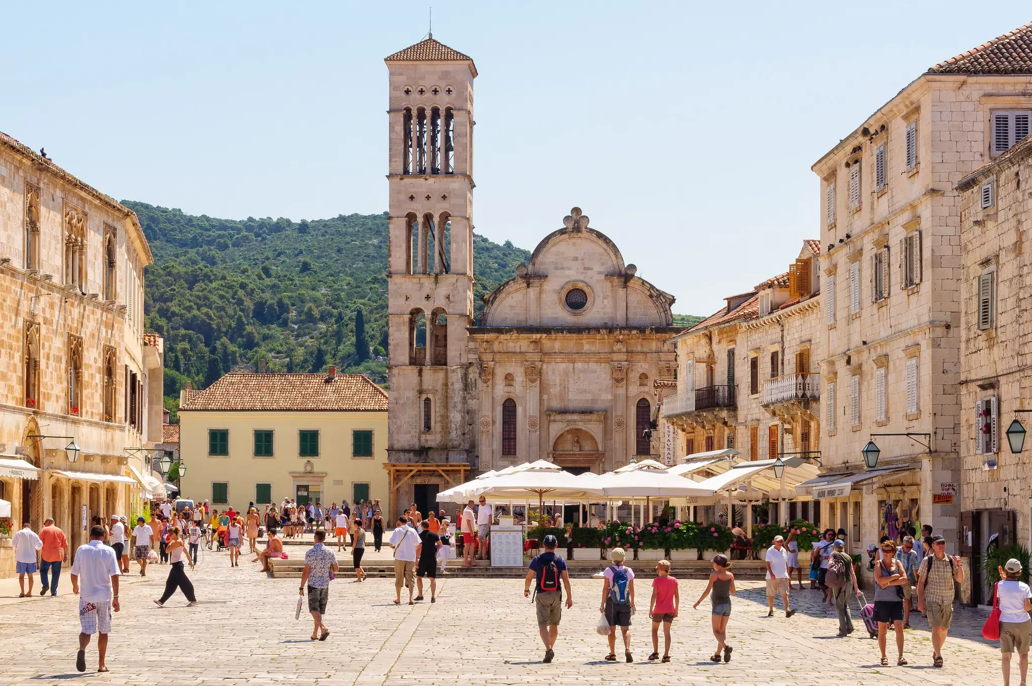People walking through a wide public square framed by a cathedral and restaurants with outdoor terraces.