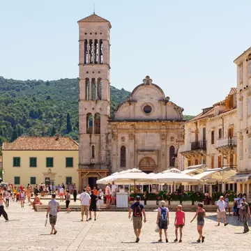 People walking through a wide public square framed by a cathedral and restaurants with outdoor terraces.