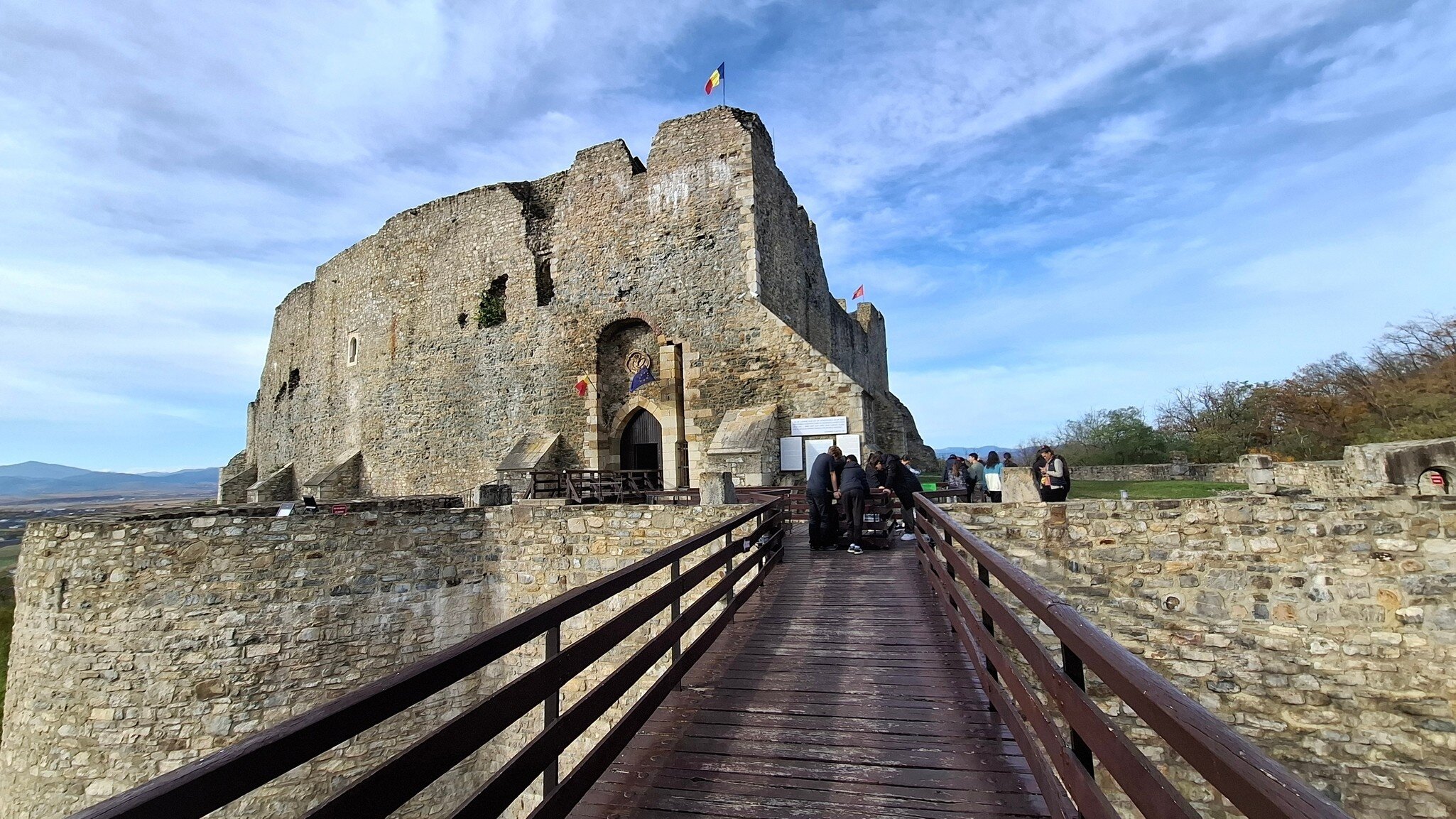 Ruined stone Neamț Citadel with Romanian flags, reached by a wooden railed bridge where a group of visitors gathers