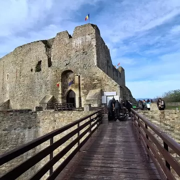 Ruined stone Neamț Citadel with Romanian flags, reached by a wooden railed bridge where a group of visitors gathers