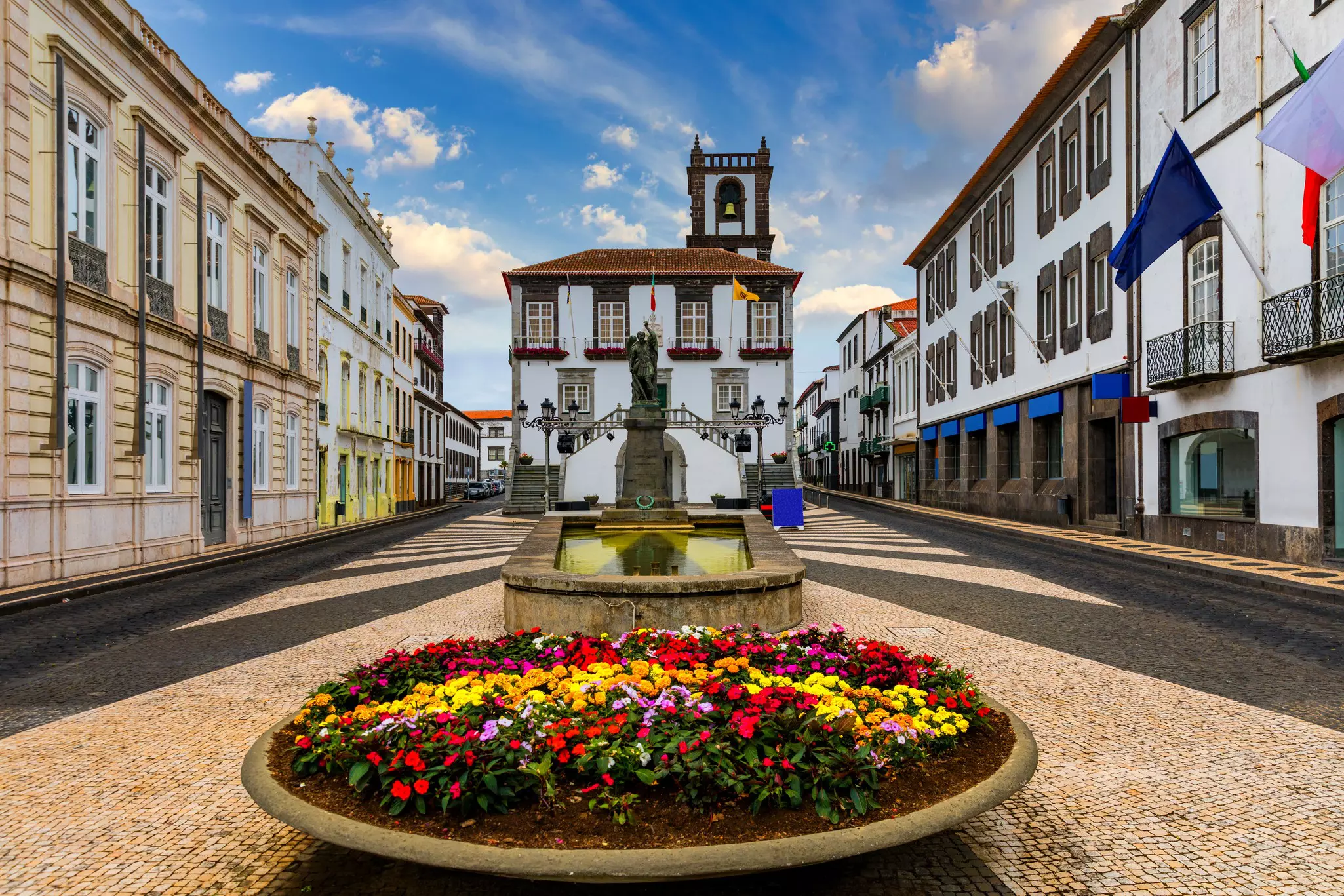 A white building at one end of an open square, with a fountain and flowerbed in the center.