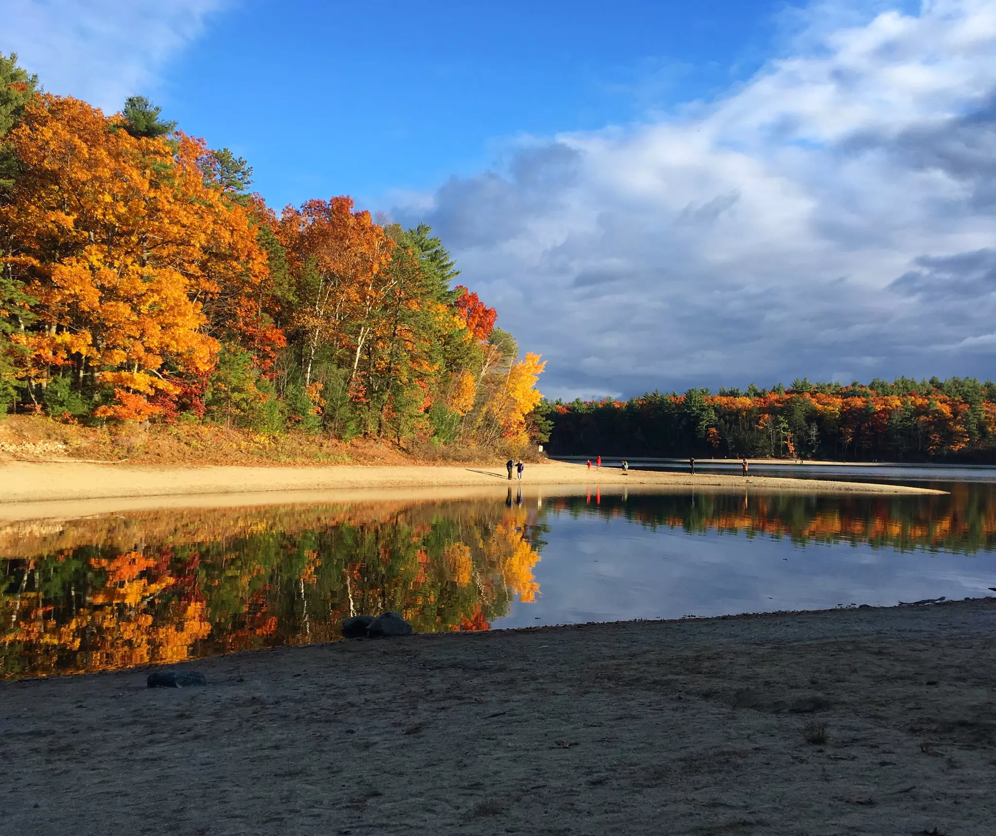 People walk on the far shore of a pond; the autumn trees are reflected in the still surface of the water.
