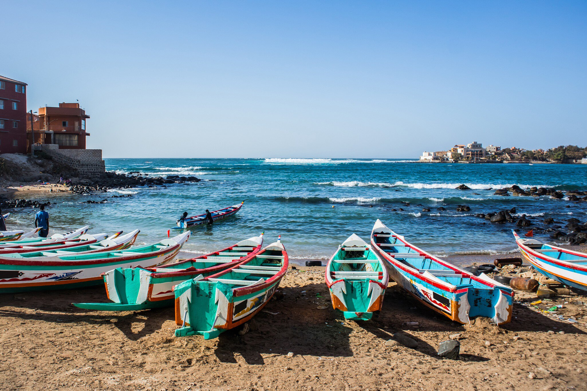 A row of narrow wooden fishing boats painted in green, red and blue pulled onto a sandy beach, surf breaking over dark rocks and a low headland beyond.