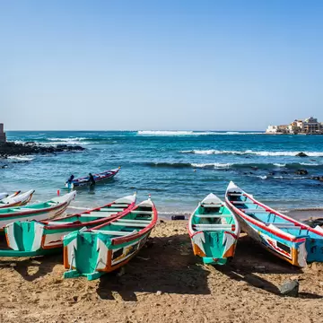 A row of narrow wooden fishing boats painted in green, red and blue pulled onto a sandy beach, surf breaking over dark rocks and a low headland beyond.
