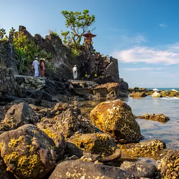 Barnacle-covered boulders in shallow water below a Hindu temple with shrines, people in white clothing, Senggigi, Lombok
