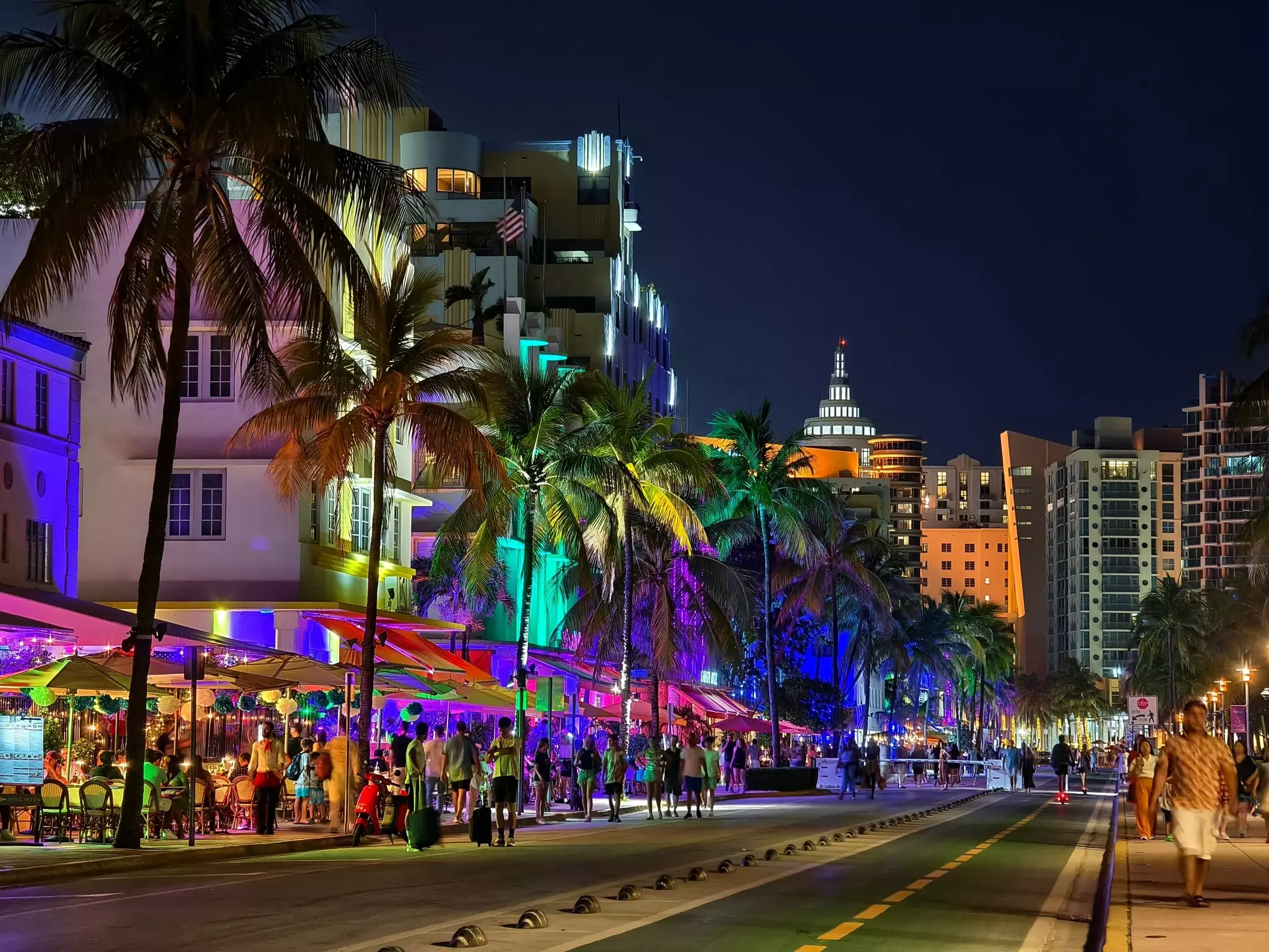 A city street is seen by night, lined with palm trees and hotels, all illuminated in colorful floodlights.