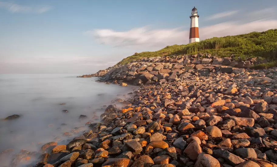 Montauk Point Lighthouse with its brown band on a grassy bluff above a rocky beach, Long Island, New York, misty sea at left