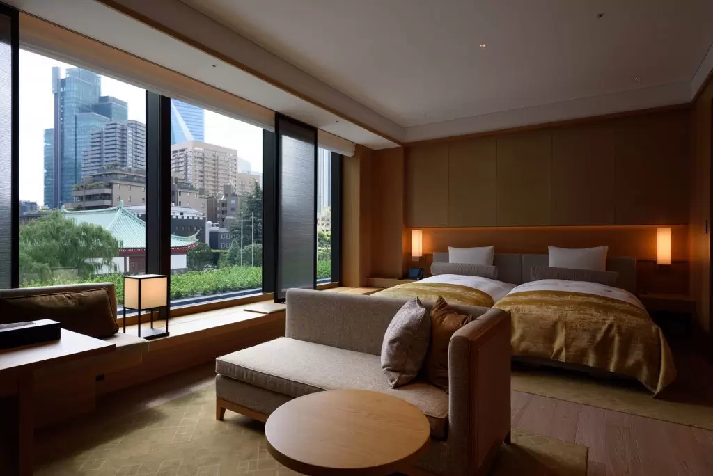 Plush, earth-toned beds and a couch in a hotel room with large windows in Tokyo