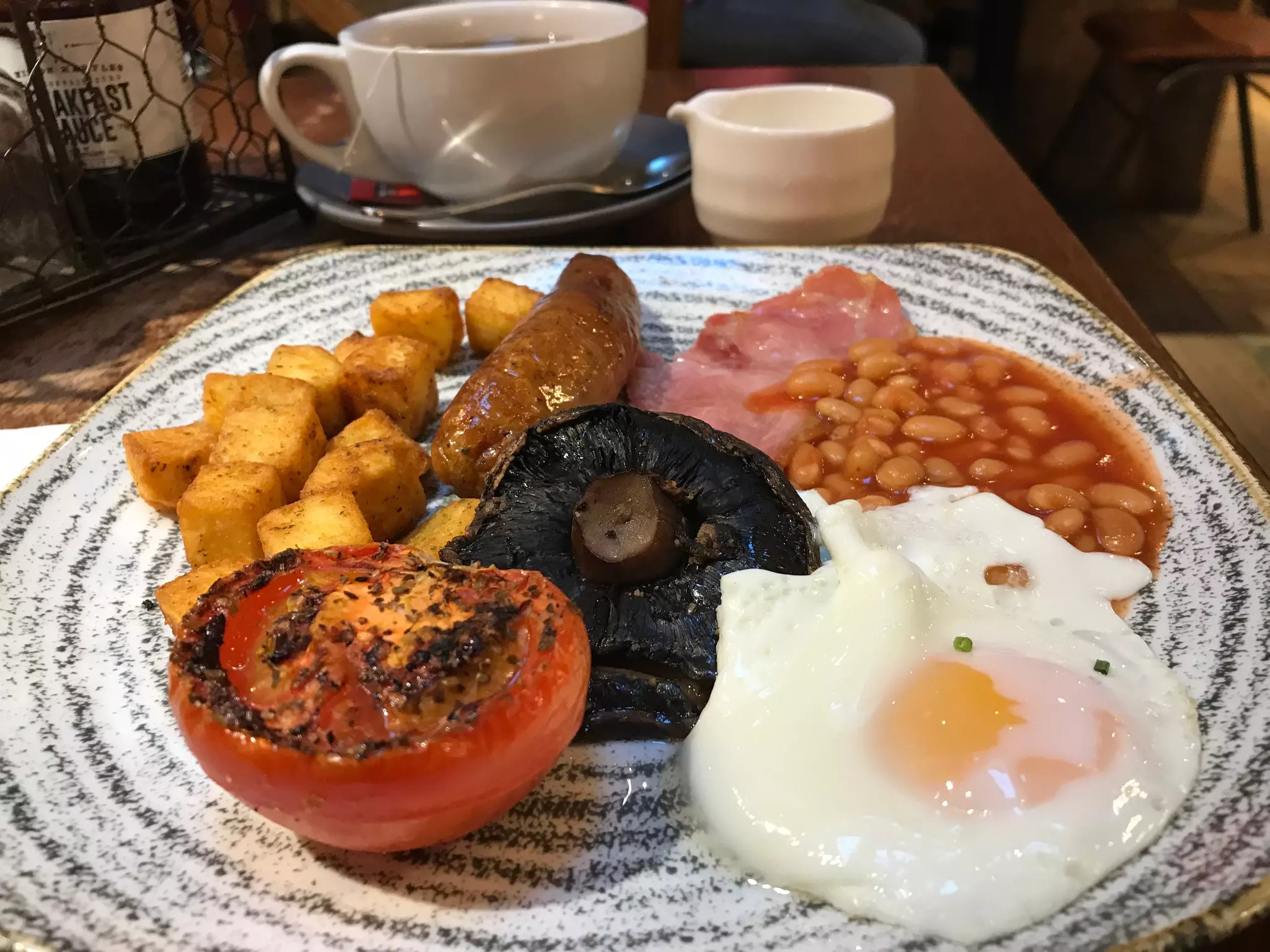 A full English breakfast at a restaurant, including bacon, sausage, egg, beans, tomato, mushroom and fried potato
