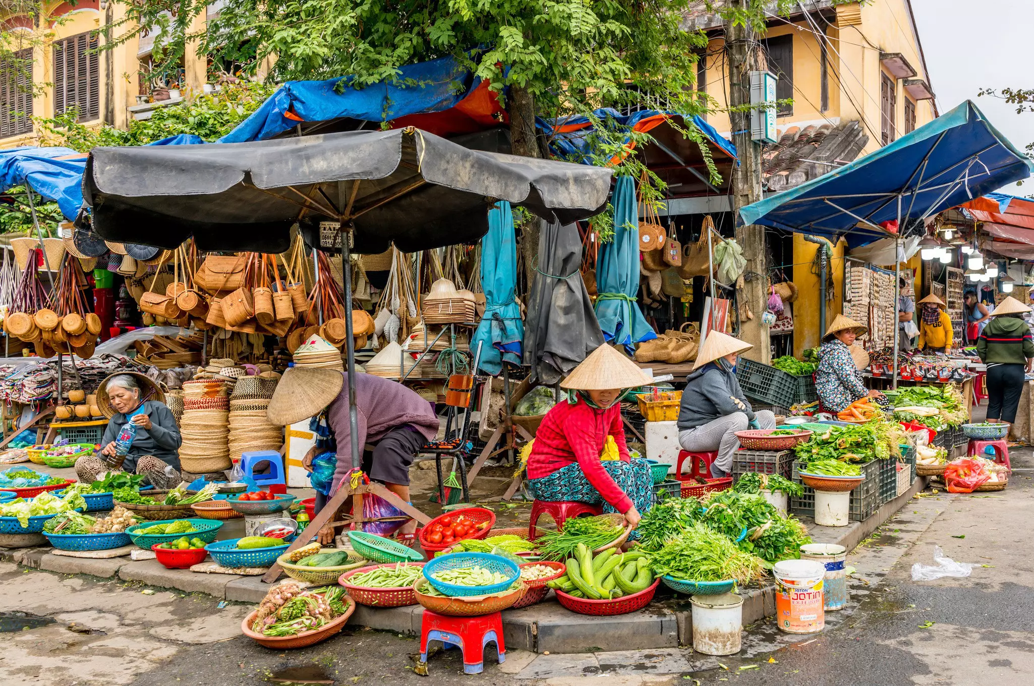 People wearing conical hats sell fresh fruits and vegetables from colorful plastic bowls lining the edge of a street.
