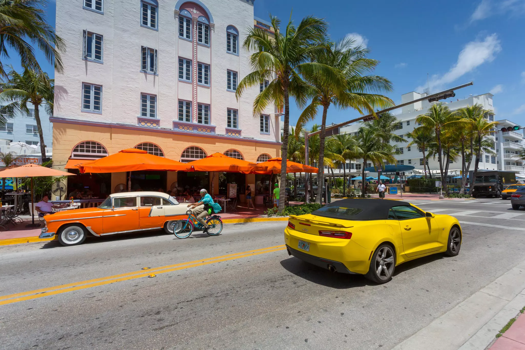 A yellow sports car goes by an orange vintage car parked on a street dotted with palm trees