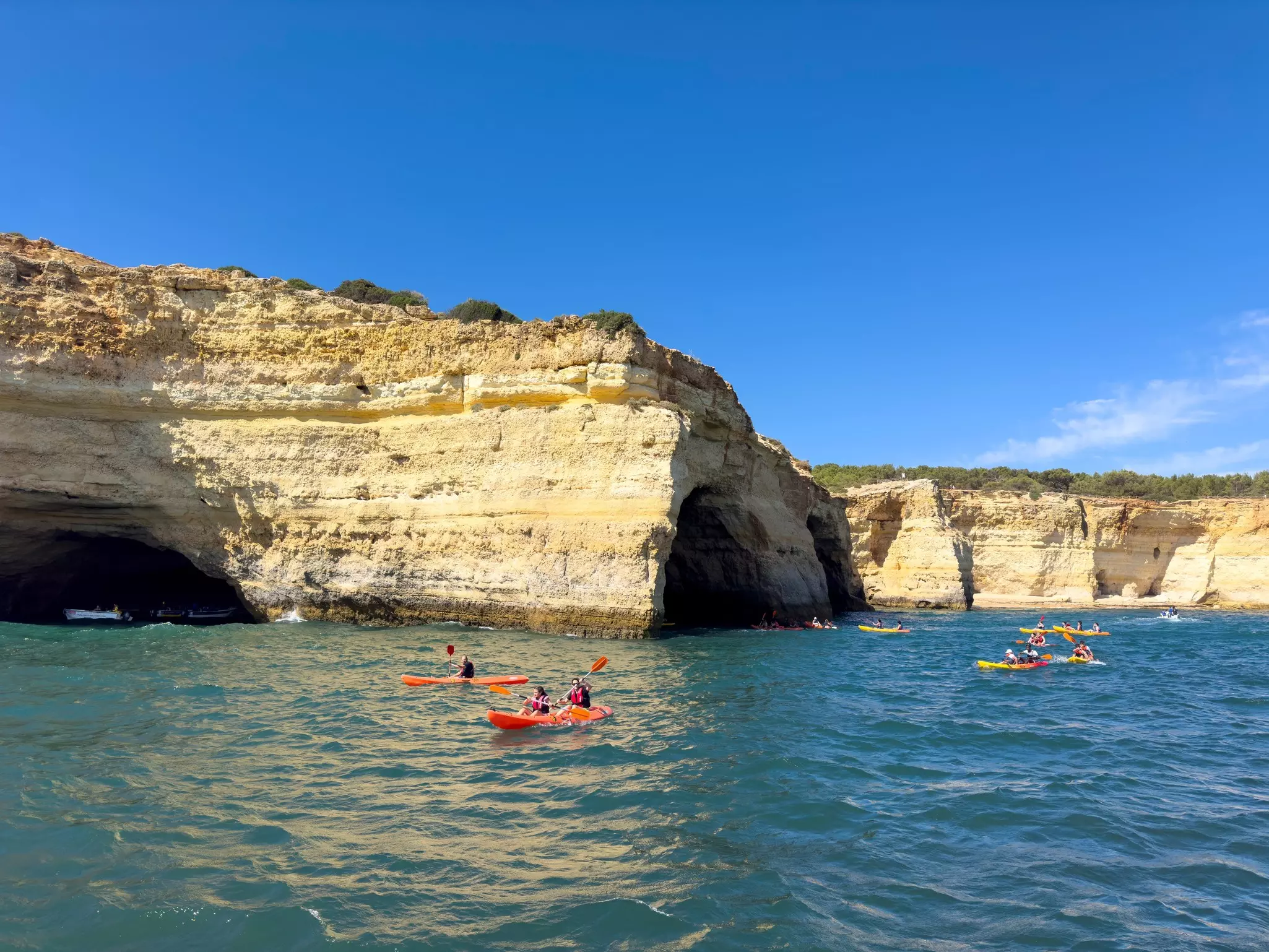 People in kayaks paddle on the sea past striated cliffs.