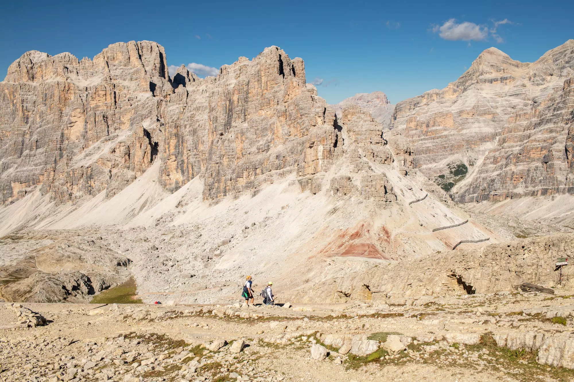 Two distant hikers against a rocky mountain landscape under blue sky in Italy.