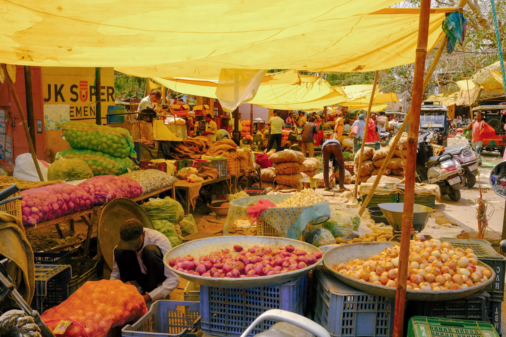 A series of vegetable stalls in a market with each of the vendors' stalls covered by yellow tarpaulins supported by large bamboo canes.