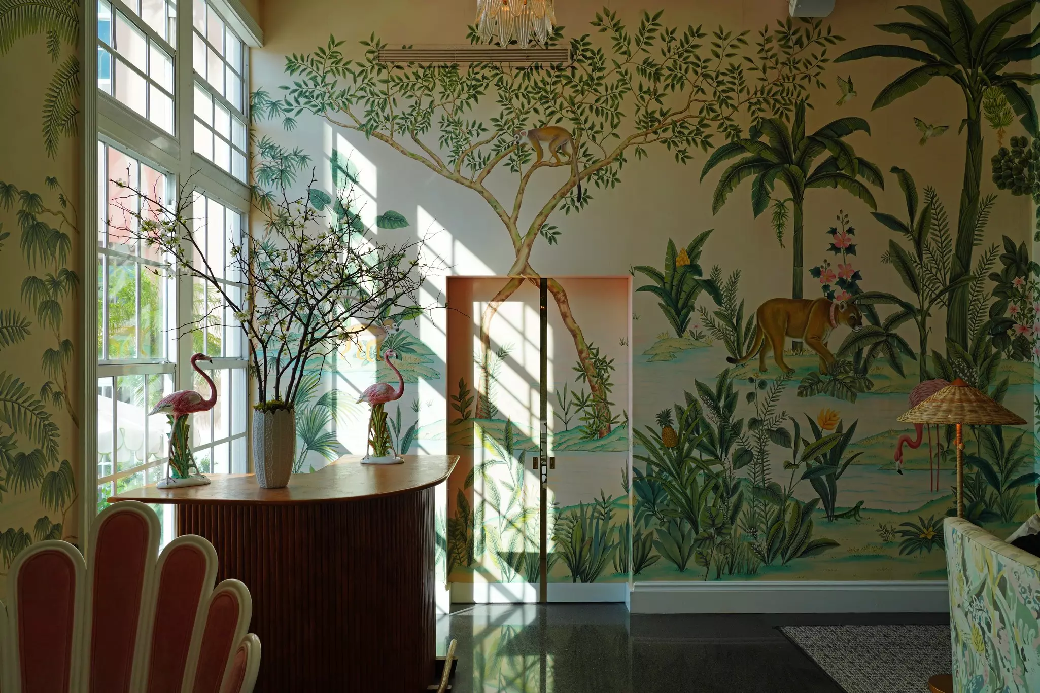 A room with a floor-to-ceiling window on one side has wallpaper with a green tropical print; there is a tall  dark wood desk with sculptures of pink flamingos.
