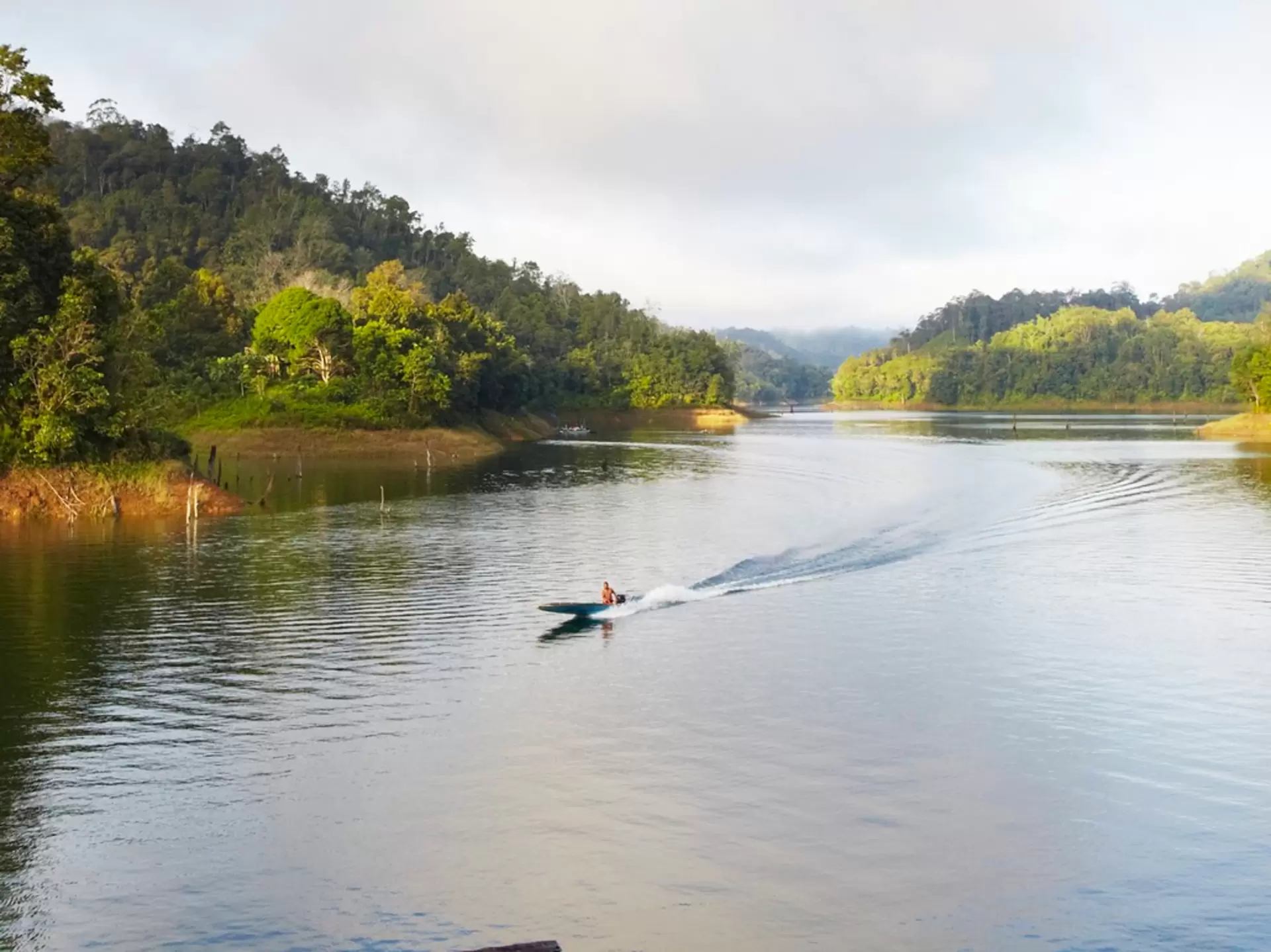A long wooden boat that's low to the water level travels down a wide river through jungle in Batang Ai National Park, Malaysia