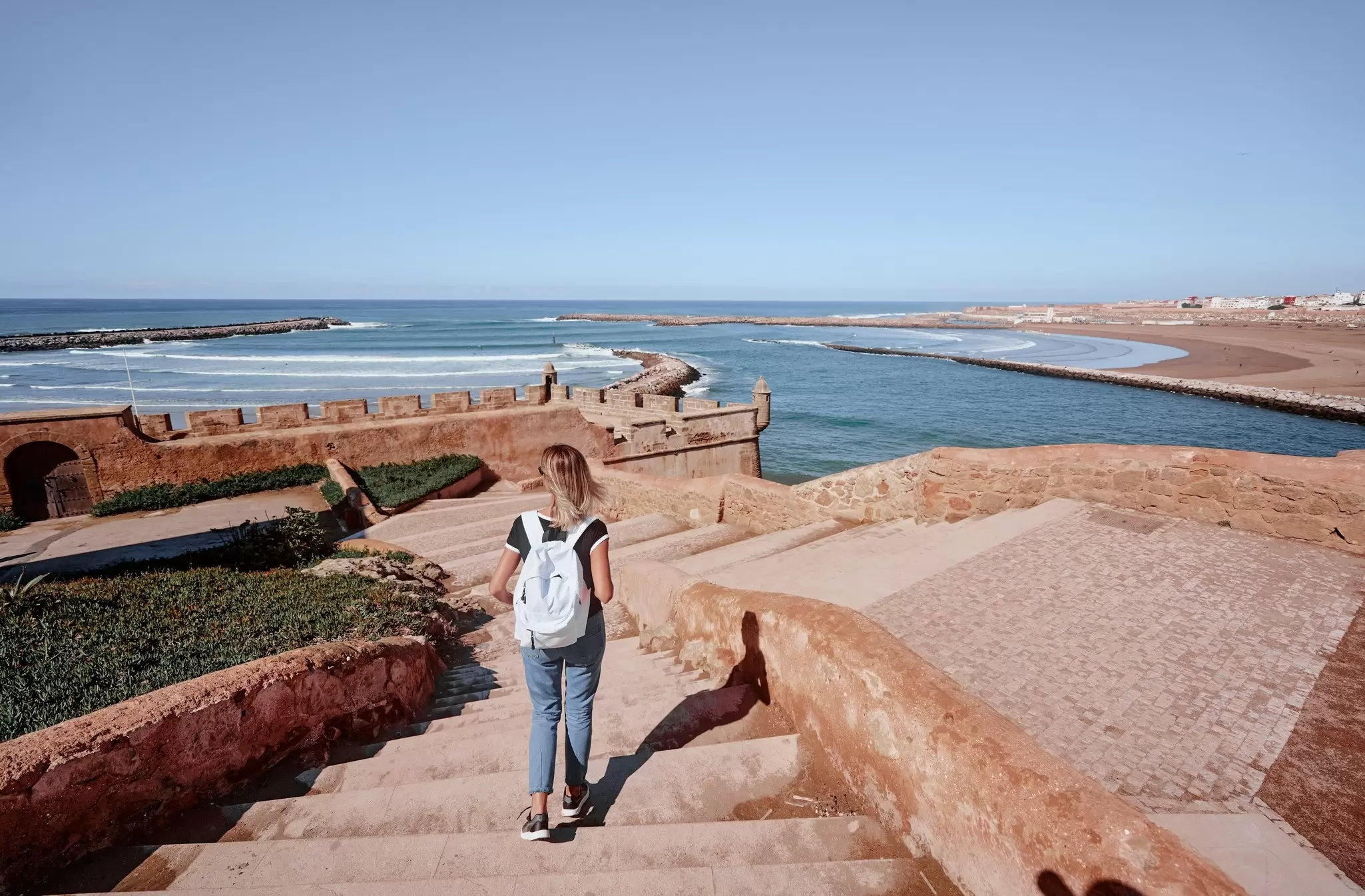 A person walking down steps at a stone fortification by the sea; a beach is in the background.