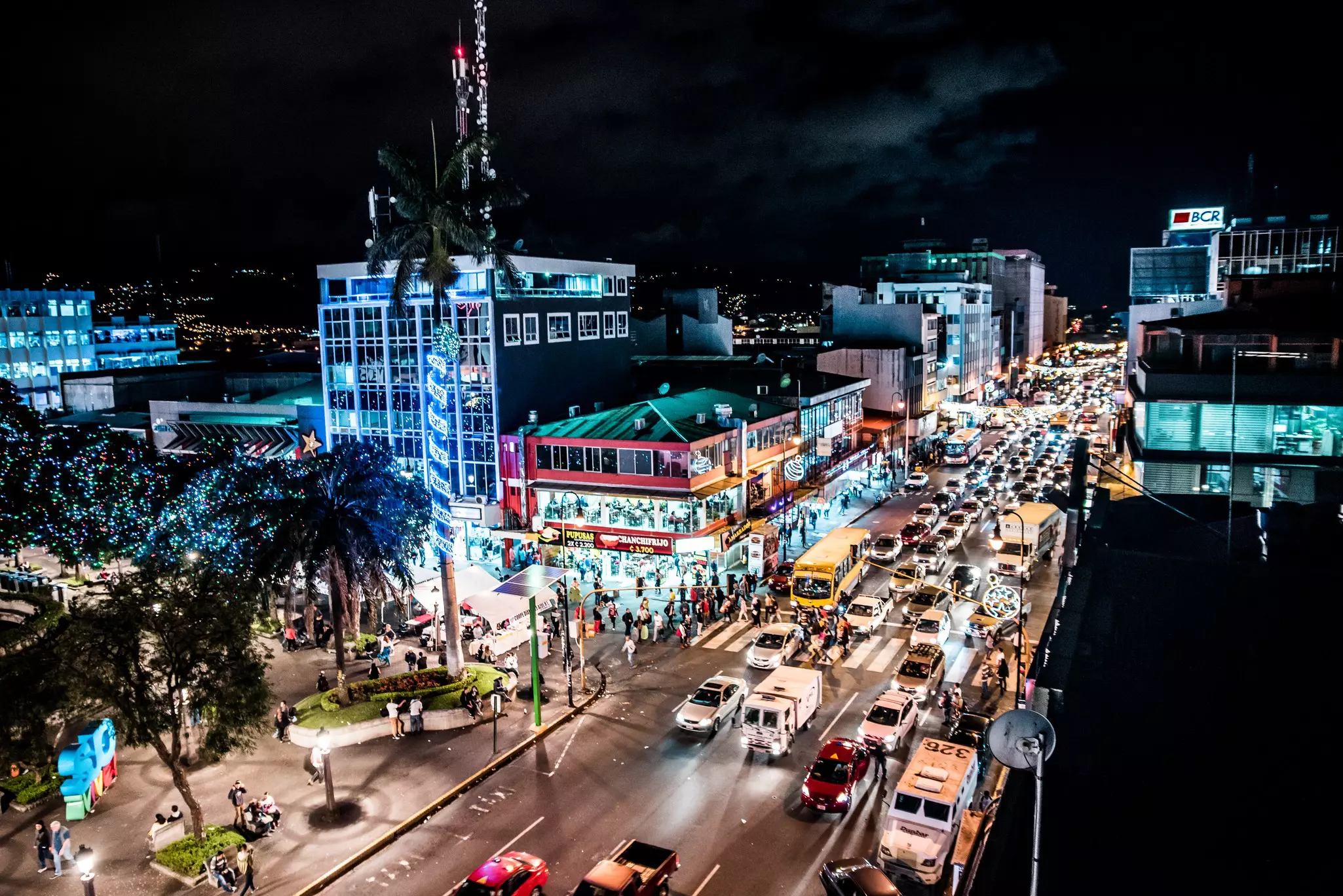 An overview of traffic on a busy street in a city at night. Illuminated buildings line the street and people walk on the sidewalks.
