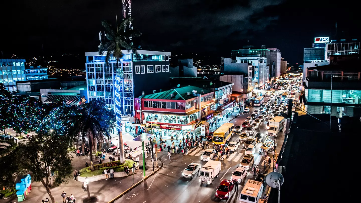 An overview of traffic on a busy street in a city at night. Illuminated buildings line the street and people walk on the sidewalks.