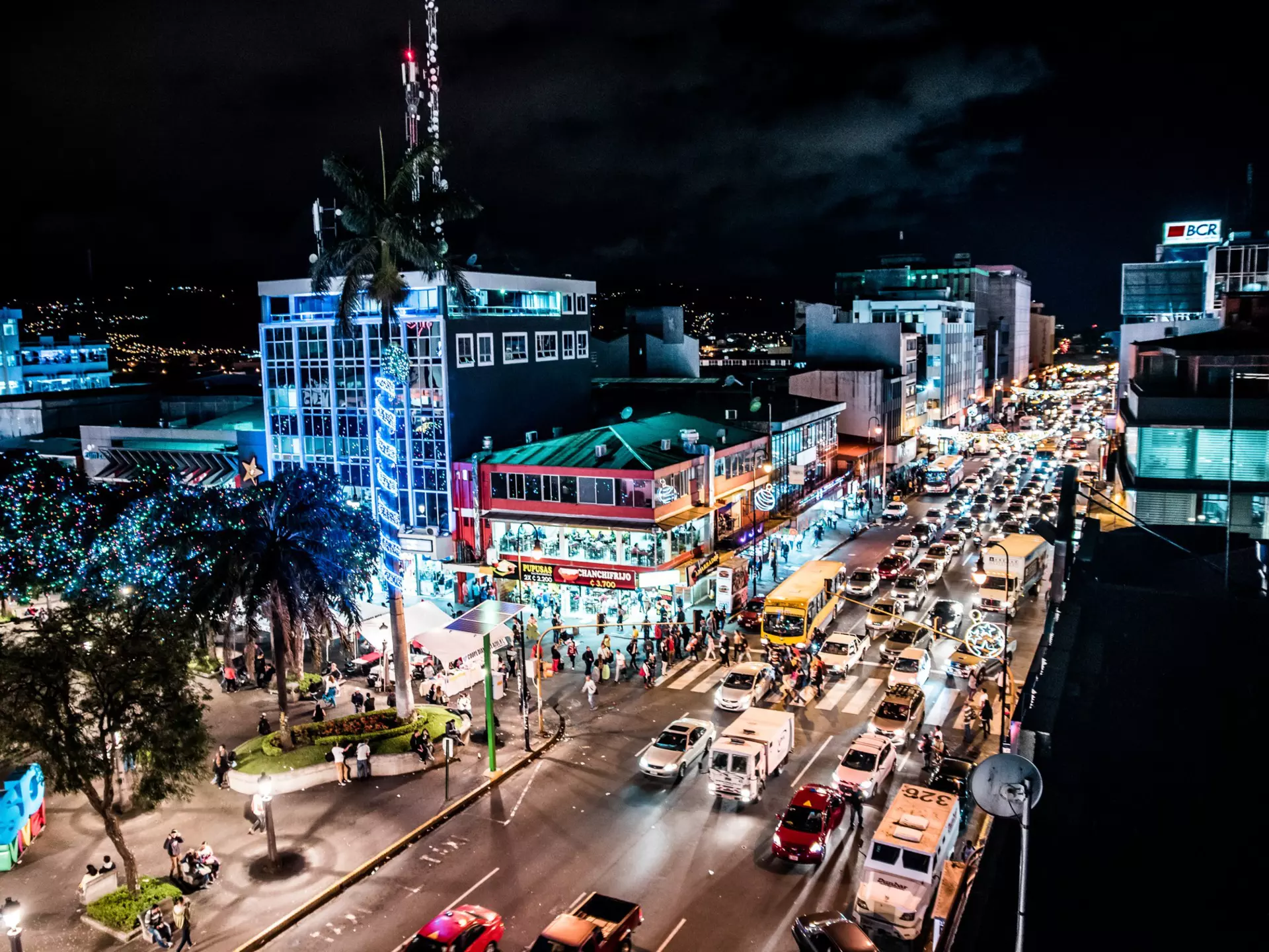 An overview of traffic on a busy street in a city at night. Illuminated buildings line the street and people walk on the sidewalks.