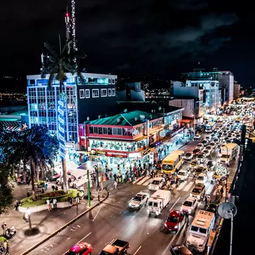 An overview of traffic on a busy street in a city at night. Illuminated buildings line the street and people walk on the sidewalks.